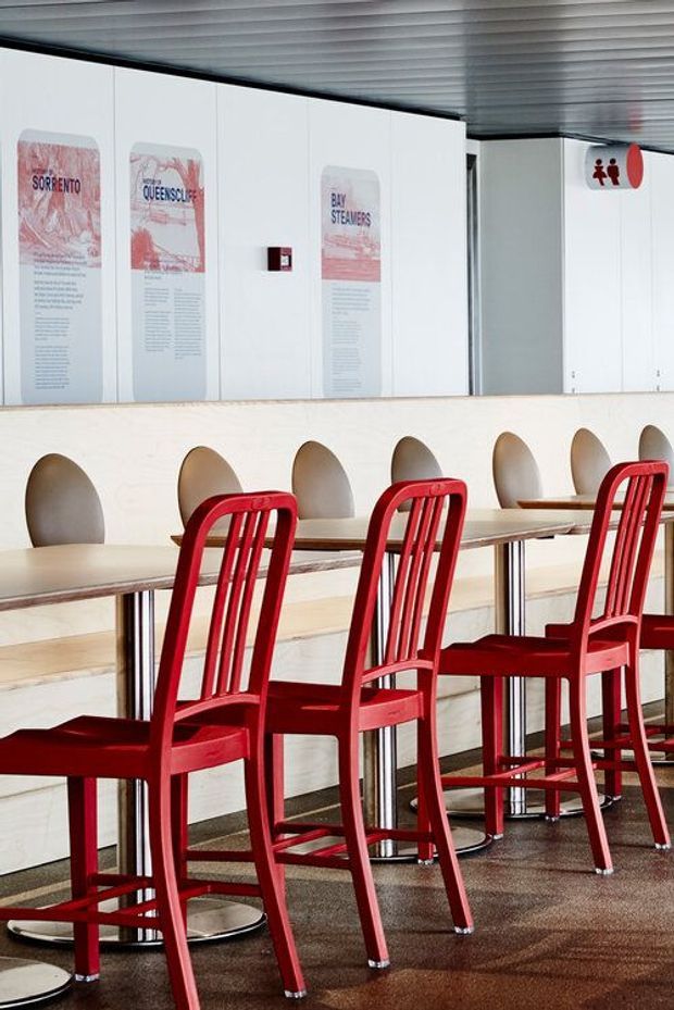 A modern commercial dining area or cafeteria with a long pale stone or concrete counter, integrated circular alcoves, and high communal tables. Bright red wooden or painted chairs line the tables. White wall panels display informational posters, while a ribbed metallic ceiling and restroom signage reinforce the institutional hospitality setting.