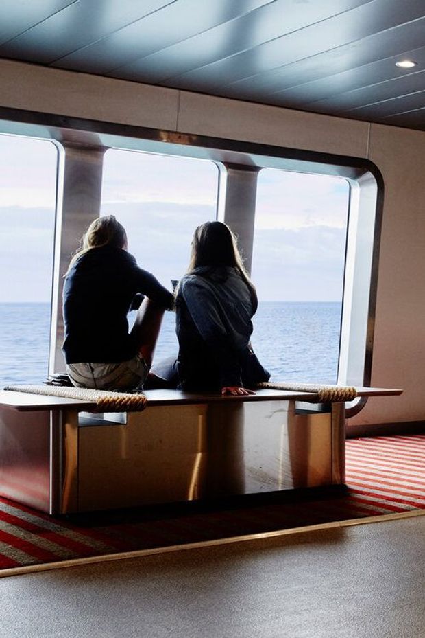 Interior of a passenger vessel with two people seated on a built-in bench beside large rounded-rectangular windows overlooking the ocean. The space has a dark paneled ceiling with recessed lights, warm wood-toned wall and joinery panels, rope detailing along the bench, and a red-and-cream striped carpet.