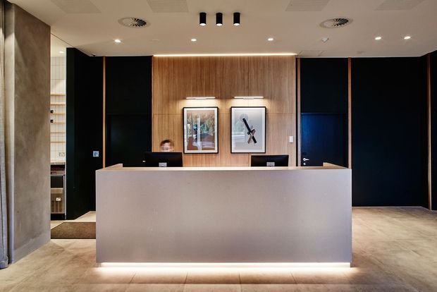 A contemporary reception area with a large pale stone or solid-surface reception desk, dark charcoal wall panels and doors, and a warm timber feature wall illuminated by concealed linear lighting. Framed artwork, recessed ceiling lights, and polished neutral flooring create a refined commercial lobby atmosphere.
