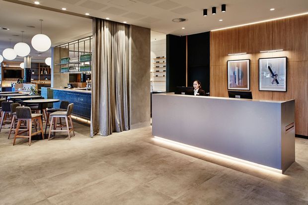A contemporary commercial reception and hospitality space featuring a large illuminated grey reception desk, warm timber wall panelling, framed artwork, recessed and pendant lighting, and an adjacent café or lounge area with tables and chairs. A curtain-lined opening and open shelving add visual separation and texture.