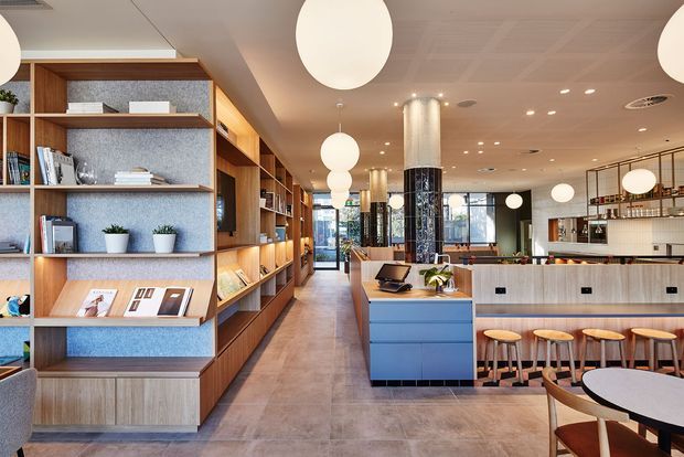 Contemporary commercial café or hospitality interior with a long built-in timber and blue shelving display on the left, a central service counter and island, and a tiled bar seating area on the right. Warm globe pendant lights, pale timber finishes, blue cabinetry, patterned tiled columns, and large windows create a bright, refined atmosphere.