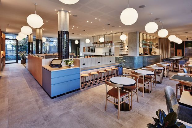 A bright contemporary hospitality interior, likely a café or restaurant, with an open bar and service counter, tiled island front, timber cabinetry, shelving, and extensive dining seating. Large spherical pendant lights hang across the ceiling, while full-height glazing and glazed doors bring in daylight. The space has a polished, welcoming fit-out with a mix of timber, blue cabinetry, patterned tile, metal shelving, and concrete-look flooring.