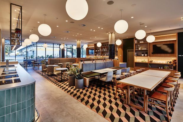 A stylish contemporary hospitality interior with an open dining and lounge layout. The space features a long communal dining table, upholstered banquette seating, timber tables and chairs, a tiled bar or service counter, extensive glazing, warm timber built-in shelving, and numerous spherical pendant lights. A bold geometric black, cream, and terracotta patterned carpet defines the central seating area.