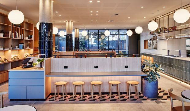 A spacious contemporary hospitality interior, likely a café, restaurant, or hotel lounge, with a long pale-wood service counter and five stools in the foreground. The space features built-in timber shelving, a tiled bar counter, dark glossy tiled columns, globe pendant lights, integrated display shelving, dining tables, and full-height glazing at the rear. The palette combines light timber, pale blue cabinetry, white surfaces, black accents, and warm ambient lighting.