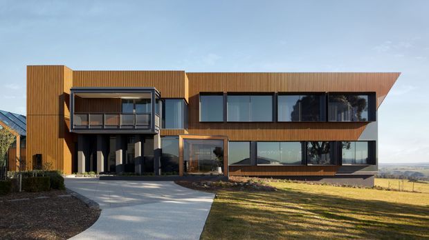 A contemporary multi-level residential house viewed from the front, set in an open rural landscape. The home features warm vertical timber cladding, extensive dark-framed glazing, projecting box-like volumes, a cantilevered upper level, and a broad asymmetrical roof plane. A curved concrete driveway approaches the entrance, with landscaped planting around the facade and open fields beyond.