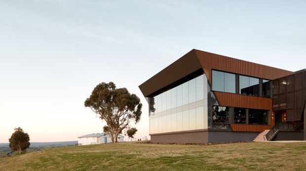 Exterior view of a contemporary multi-level building set on an open grassy, gently sloping site. The building features strong angular roof and facade forms, extensive floor-to-ceiling glazing, dark metal framing, and warm rust-brown vertical metal cladding. Exterior stairs lead to an elevated entrance, with a broad rural or semi-rural horizon visible beyond.