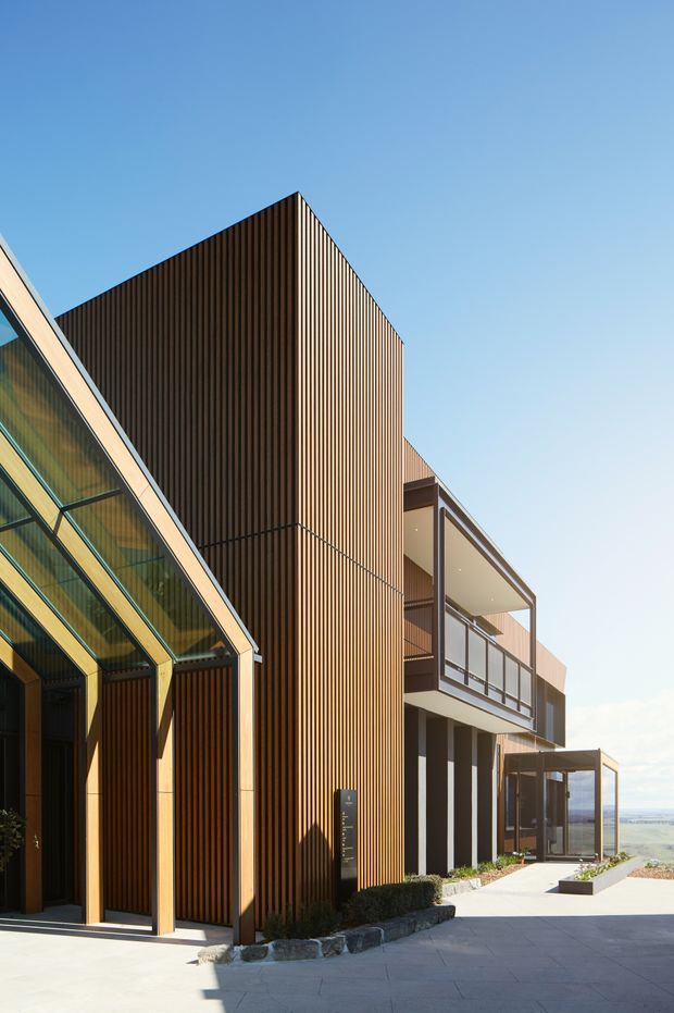 Exterior architectural photograph of a contemporary multi-level building on a coastal or elevated site. The facade combines strong vertical timber-look slatted cladding with dark framing, extensive glazing, projecting balconies, and a steeply angled glazed volume at the left. A paved forecourt and planted edge sit in front, with distant water or coastline visible beyond the building.