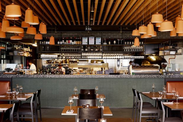 A warmly lit contemporary restaurant interior viewed from the dining area toward a long bar and open service counter. The space features a dark green small-format tiled bar front, dark shelving filled with bottles and glassware, timber ceiling battens, black ceiling structure, abundant pendant lights, timber dining tables, and dark upholstered dining chairs. Staff, bar equipment, and a large metallic cooking hood are visible behind the counter.