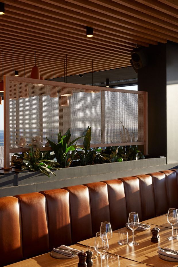 A warmly lit contemporary restaurant dining area with a long upholstered brown leather banquette, timber dining tables set with glassware and table settings, and a raised planter dividing the seating from a perforated timber privacy screen. The ceiling features closely spaced exposed timber battens with suspended pendant lights, while dark structural columns and indoor planting add contrast. The space has a refined hospitality interior with strong natural materials and layered lighting.