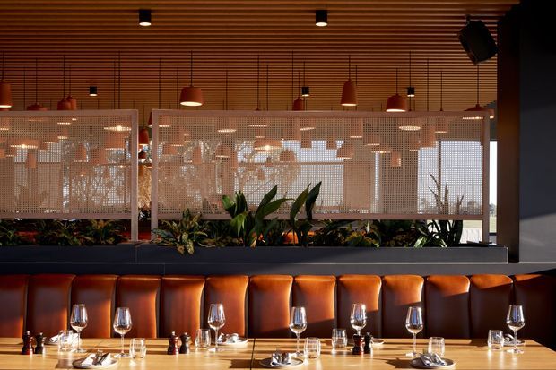 A warmly lit contemporary restaurant dining area with a long upholstered banquette in rich caramel leather, set with white tablecloth-free tables, wine glasses, plates, and cutlery. A raised planter filled with lush greenery separates the seating from a perforated metal privacy screen and windows. The ceiling features extensive linear timber slats with recessed downlights and suspended pendant lights, creating a refined hospitality interior.