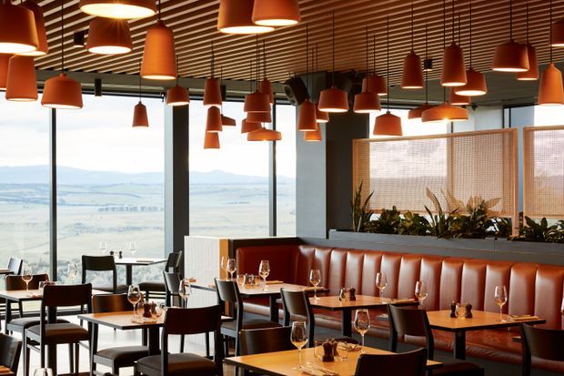 A contemporary restaurant dining area with extensive floor-to-ceiling glazing overlooking a broad landscape. The space features many suspended terracotta pendant lights, a slatted timber ceiling, dark structural columns, warm timber dining tables, dark chairs, and a long cognac leather banquette with integrated planter screening.