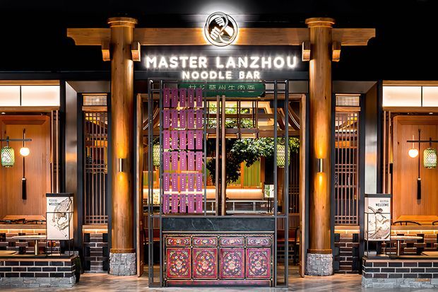 Front-facing view of the entrance to Master Lanzhou Noodle Bar. The commercial restaurant facade features illuminated signage, substantial timber posts, dark metal grilles, warm wood wall panels, patterned decorative panels on the host counter, brickwork along the lower walls, and warm interior lighting. Dining areas are visible beyond the entrance.