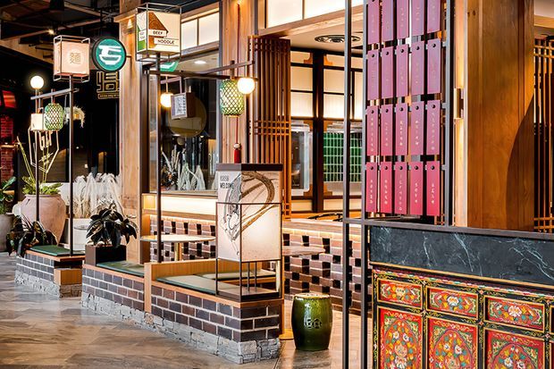 A richly detailed commercial interior, likely a restaurant or hospitality venue, featuring timber-framed glazed partitions, dark metal framing, exposed ceiling services, decorative pendant lights, tiled and brick-clad low partitions, indoor planting, and brightly patterned decorative wall panels. The space has an eclectic Asian-inspired character with layered materials, strong colour, and reflective glazing.