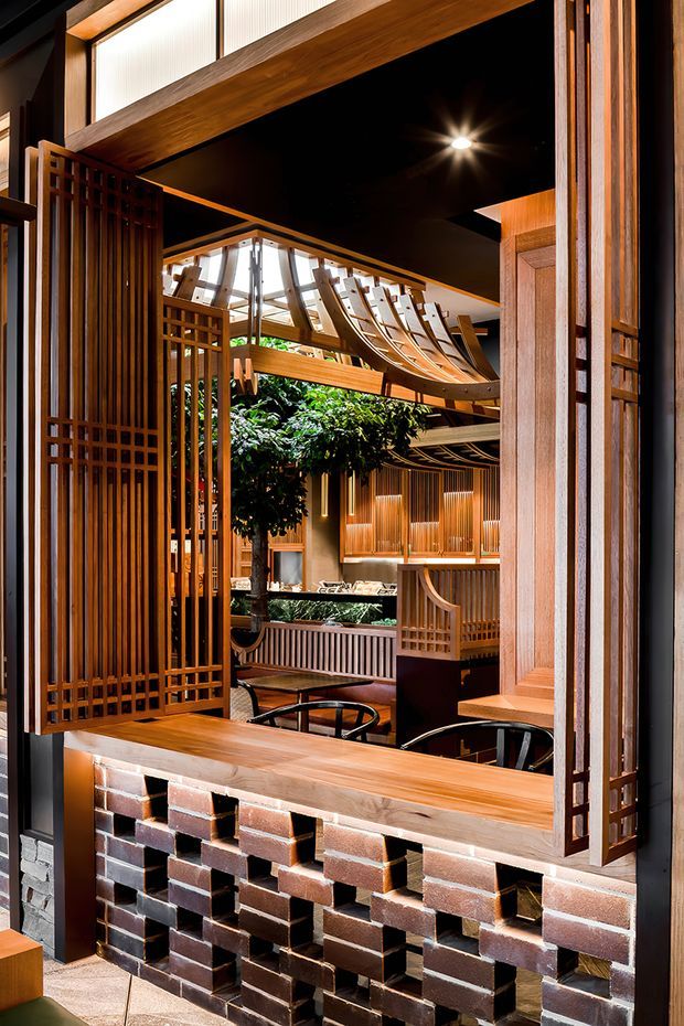 A richly detailed interior view through a timber-framed opening, featuring dark stained wood screens, vertical slatted joinery, exposed brickwork, warm timber surfaces, and a dramatic black ceiling. The space beyond appears to be a multi-level hospitality or commercial interior with indoor planting, sculptural suspended timber elements, railings, and seating. The composition highlights layered screens, custom woodwork, and warm ambient lighting.