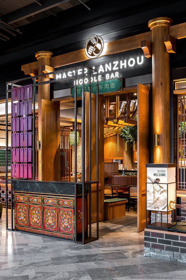 A richly detailed entrance to a Chinese-style restaurant and noodle bar, with prominent timber columns and beams, black metal framing, illuminated signage, decorative red and turquoise carved panels, and warm timber-lined seating visible inside. The commercial frontage combines traditional Asian-inspired ornament with an industrial loft-like ceiling and dark structural elements.