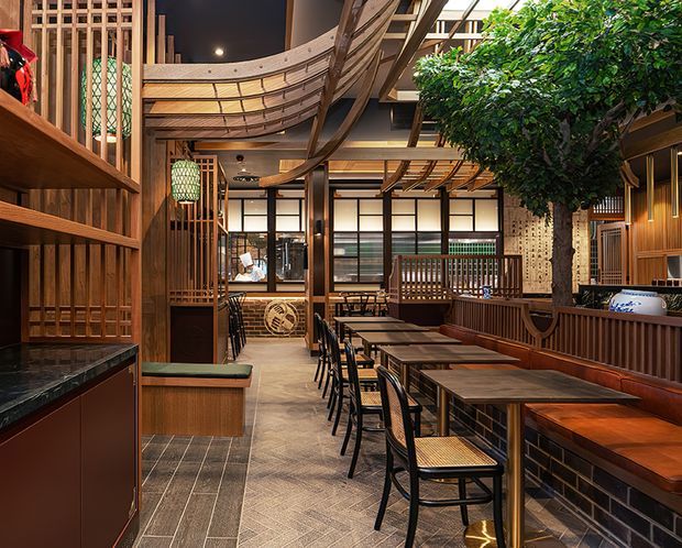 A richly detailed Japanese-inspired restaurant interior with rows of dark timber dining tables and black metal chairs, built-in banquette seating, timber lattice screens, framed glazing, brick-faced counters, decorative lanterns, and a prominent indoor tree. Curved timber ceiling elements and layered joinery create a warm, crafted hospitality setting.