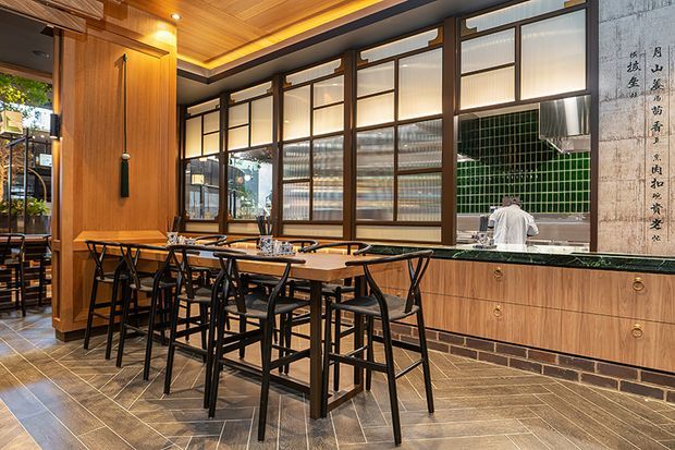 A warmly lit restaurant or hospitality dining area with a long communal timber table and black-framed stools and chairs. The space features extensive dark timber-framed glazing, a timber slatted ceiling with concealed lighting, wood-panelled cabinetry, a dark green tiled wall, exposed brick detailing, and decorative calligraphy panels. A service or kitchen area is visible beyond the glazing.