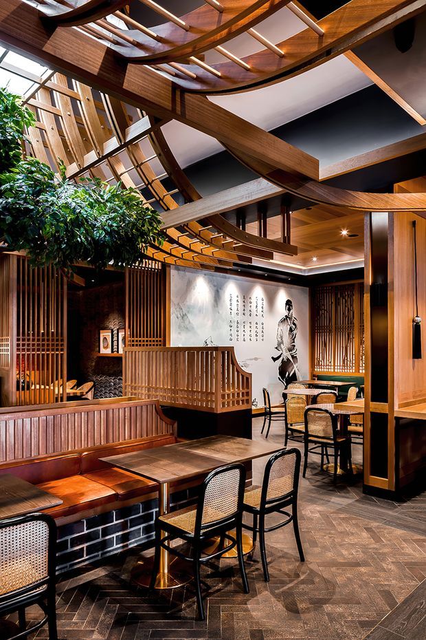 A richly detailed Japanese-inspired restaurant interior with dining tables, woven-seat chairs, leather banquette seating, timber screens, slatted partitions, and a dramatic sculptural timber ceiling with skylights. Dark patterned floor tiles, warm wood finishes, black accents, indoor planting, and a large monochrome mural create a refined, atmospheric hospitality space.