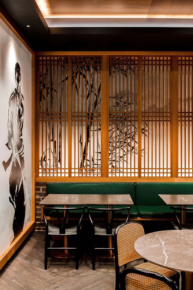 A warmly lit restaurant or hospitality dining area with green upholstered banquette seating, small marble-topped tables, black-and-cane chairs, and a striking timber lattice screen backed by botanical imagery. The space features dark timber flooring, timber ceiling panelling, black ceiling sections, integrated linear lighting, and monochrome wall artwork, creating a refined Japanese-inspired contemporary interior.