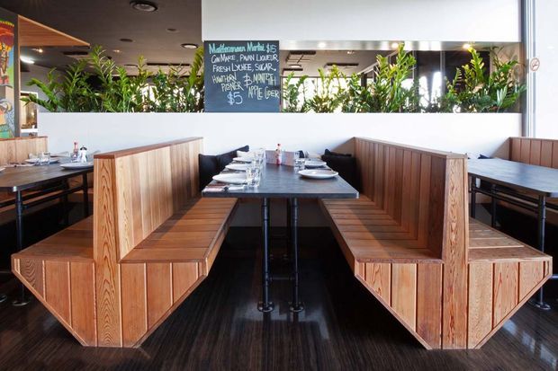 Interior photograph of a contemporary casual restaurant dining area with dark tables, built-in timber booth seating, angular timber partitions, black metal table bases, and a long mirrored planter partition filled with greenery. A handwritten menu board is visible above the booths. The space has a warm timber-and-black palette and integrated banquette seating.