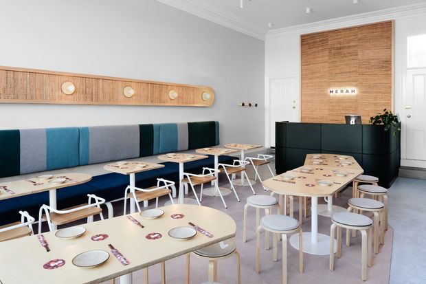 Bright contemporary café or casual dining interior with multiple light-wood tables, pale stools, and white metal-framed chairs arranged in rows. A long built-in upholstered banquette in blue, teal, and grey runs along the left wall beneath a sculptural timber wall feature with circular details. At the rear is a dark reception or service counter, framed by a large vertical timber panel bearing the word “MECHA”. The room has pale grey walls, white decorative ceiling mouldings, and a light neutral floor.
