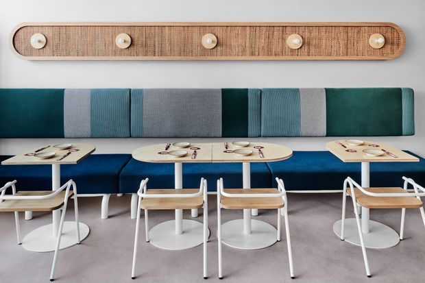 Contemporary commercial dining or hospitality space with a long built-in upholstered banquette in alternating teal and grey panels, paired with small light-wood tables and white metal-framed chairs. A horizontal light timber wall feature with circular accents spans the wall above the seating. The room has a polished, coordinated palette of teal, blue, grey, white and timber, with a clean modern fit-out.