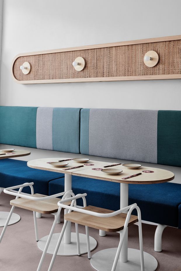 A softly lit contemporary commercial dining space with a long upholstered banquette in teal, blue, and grey panels, paired with pale wood oval tables and white pedestal bases. White tubular-frame dining chairs with timber seats and backs sit around tables set with plates and cutlery. A large rounded rectangular woven wall panel with integrated circular lights forms a feature backdrop.