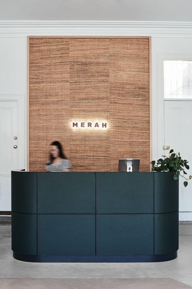A refined hotel or commercial reception area featuring a dark teal curved reception desk with integrated panel divisions and a dark plinth. Behind it is a large rectangular woven natural-fibre feature wall with illuminated MERAH lettering. The surrounding space has pale grey-white traditional wall panelling, decorative mouldings, panelled doors, and a glazed transom window. A computer terminal and leafy plant sit on the desk, with a blurred receptionist behind it.