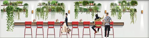 A bright contemporary commercial interior featuring a long timber counter with red metal stools, set against a white wall decorated with extensive built-in vertical gardens and potted trailing plants. Several people occupy the counter area, suggesting a café, dining, or shared workspace setting.