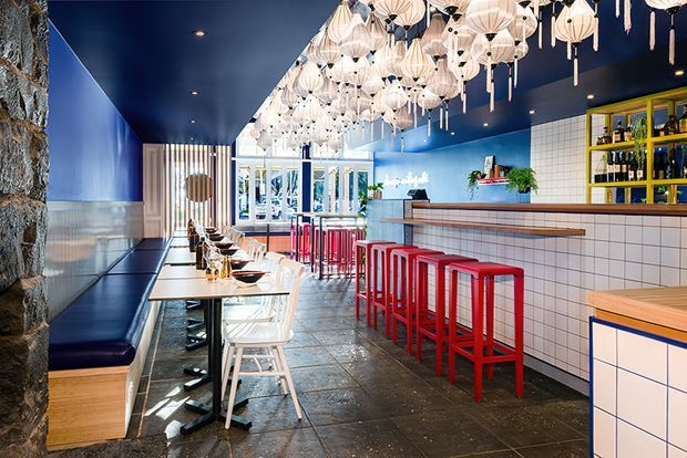A vibrant contemporary commercial dining and bar interior with a long tiled service counter, bright red bar stools, white stools and tables, and deep blue walls and ceiling. A large cluster of sculptural cream pendant lights creates a dramatic focal point above the counter and seating. The space includes a dark built-in banquette along the left wall, open shelving with bottles behind the counter, patterned floor tiles, and a bright glazed frontage at the rear.