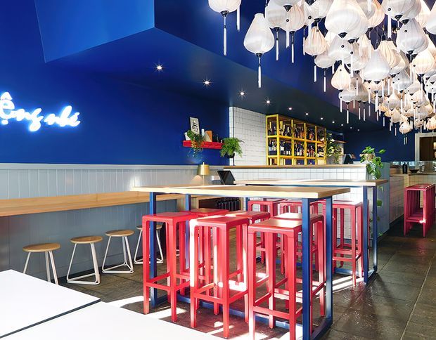 Vibrant commercial bar or café interior with deep blue walls and ceiling, a large cluster of white pendant lights, communal timber-topped high tables, bright red bar stools, a long built-in bench, and illuminated shelving behind the counter. The space has a bold, playful hospitality design with strong colour contrast and tiled and dark flooring.