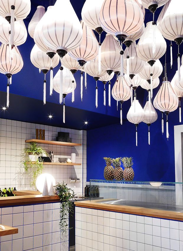A bold, highly stylised commercial or hospitality interior with vivid cobalt-blue walls and ceiling, white square wall tiles, warm timber shelving and counters, and numerous suspended translucent lantern pendants. A tiled service counter with glass display glazing occupies the foreground, with decorative pineapples and plants adding visual interest. The dramatic lighting and saturated palette create an ornate, playful atmosphere.
