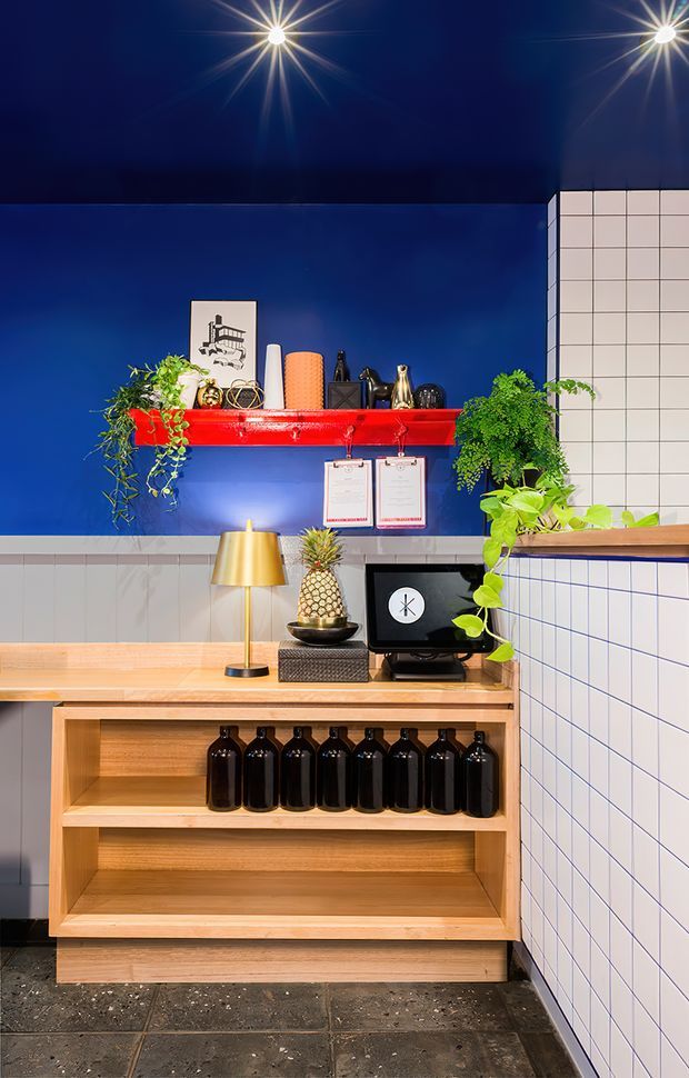 A vividly styled commercial counter or bar area with deep cobalt-blue walls, a glossy red wall shelf, white square-tile wainscoting and partition walls, and a warm timber built-in counter with open display shelving. Black bottles, plants, artwork, a gold table lamp and a computer sit on the counter, while dark tiled flooring and dramatic starburst ceiling lights reinforce the bold, eclectic fit-out.