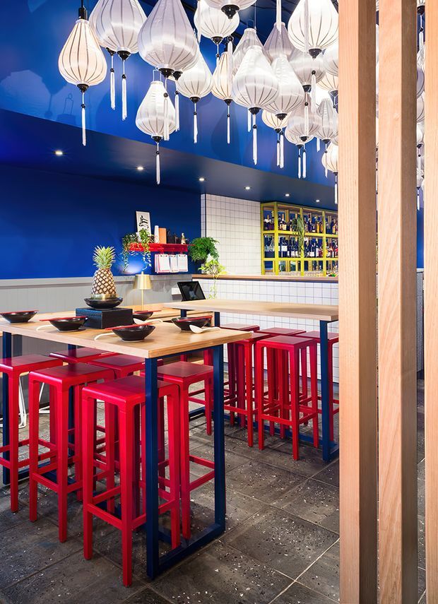 A vibrant restaurant dining area with deep cobalt-blue walls and ceiling, white tiled wall sections, and a bar counter with yellow-framed bottle shelving. Light timber communal tables are paired with vivid red bar stools and dark blue metal table frames. Numerous white, lantern-like pendant lights create a dense decorative ceiling installation, while timber vertical screens frame the foreground. The space has a lively, bold, eclectic hospitality interior.