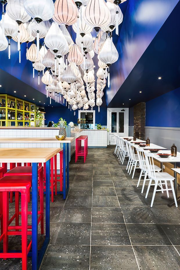 A vibrant commercial dining venue with a long bar and high communal tables. The space features deep blue walls and ceiling, white tiled bar and wall finishes, dark stone-look floor tiles, bright red and blue stools, white dining chairs, and a striking installation of numerous white and pale pink lantern pendant lights suspended overhead. Open shelving behind the bar displays bottles, while the dining area extends along the right side.