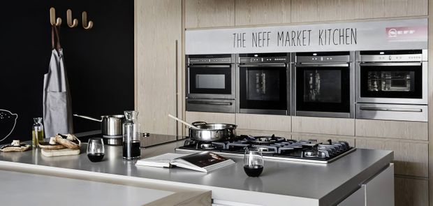 Contemporary kitchen showroom or residential kitchen featuring a large grey island with integrated gas cooktop, multiple built-in black ovens, light timber-look cabinetry, and a black accent wall with coat hooks. Stainless steel cookware and small accessories sit on the benchtop. A sign reading “THE NEFF MARKET KITCHEN” is visible above the appliances.