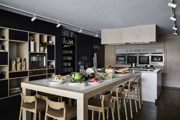 A contemporary kitchen and dining space centred on a large island with seating for several people. The room features dark charcoal walls, light timber cabinetry and shelving, integrated stainless-steel appliances, track lighting, and a substantial pale stone or composite island. Open shelving displays kitchenware, while fresh produce and serving dishes are arranged across the island.