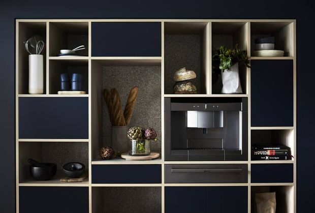 A dark, refined built-in kitchen or butler's pantry wall featuring a full-height grid of navy cabinetry and open display niches. The composition includes an integrated coffee machine, drawers, stone-textured niche back panels, open shelves with crockery and cookware, and styled food-storage displays. Warm beige framing and subdued lighting create a high-contrast, sophisticated look.