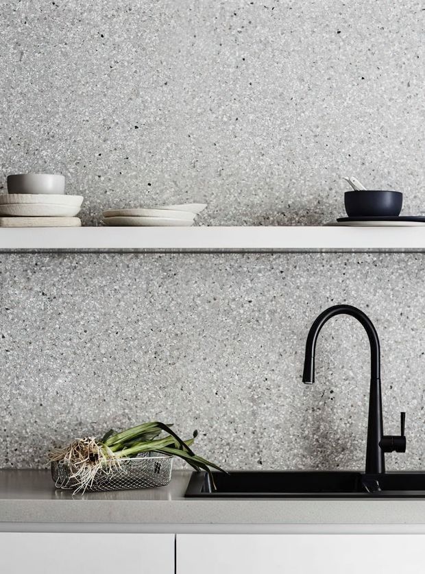 Close-up of a minimalist kitchen sink area with a black gooseneck tap, dark undermount sink, pale cabinetry, and a white open shelf holding stacked dishes and cups. The backsplash and wall are finished in a grey speckled terrazzo-like surface, creating a monochromatic contemporary composition.
