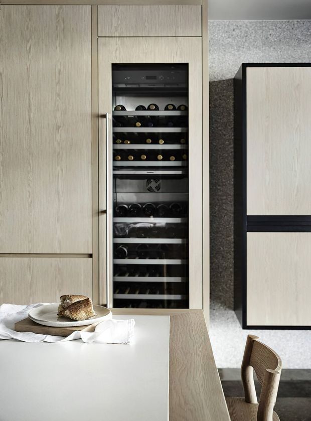 Contemporary kitchen interior featuring a tall integrated wine refrigerator set within pale timber cabinetry. A light stone or composite island benchtop occupies the foreground, with a timber dining extension and a curved timber chair. The cabinetry is paired with a dark textured stone backsplash and dark trim, creating a refined, neutral-toned kitchen fit-out.