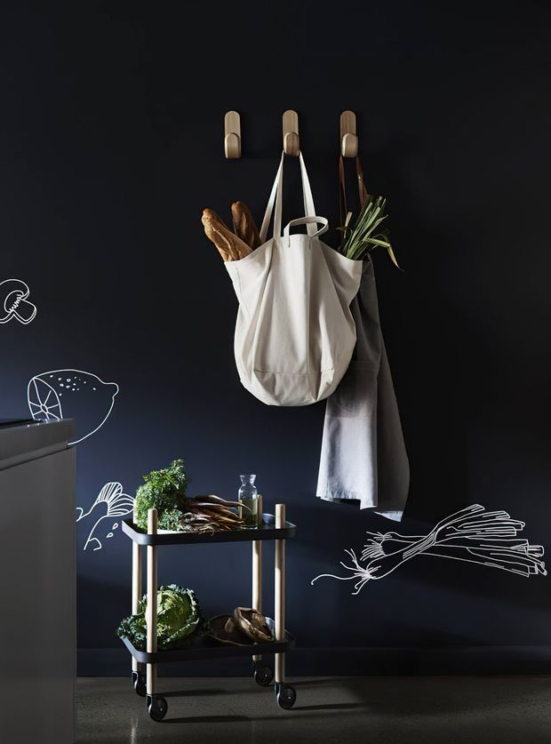 A styled interior vignette featuring a compact black rolling utility cart with timber-toned vertical supports, potted greenery, produce, bottles and kitchen items. Above it, a dark wall has three timber hooks holding a white canvas tote, a grey apron or cloth, and a smaller dark bag. White hand-drawn food and botanical illustrations decorate the wall, creating a playful, graphic kitchen or pantry-adjacent storage scene.