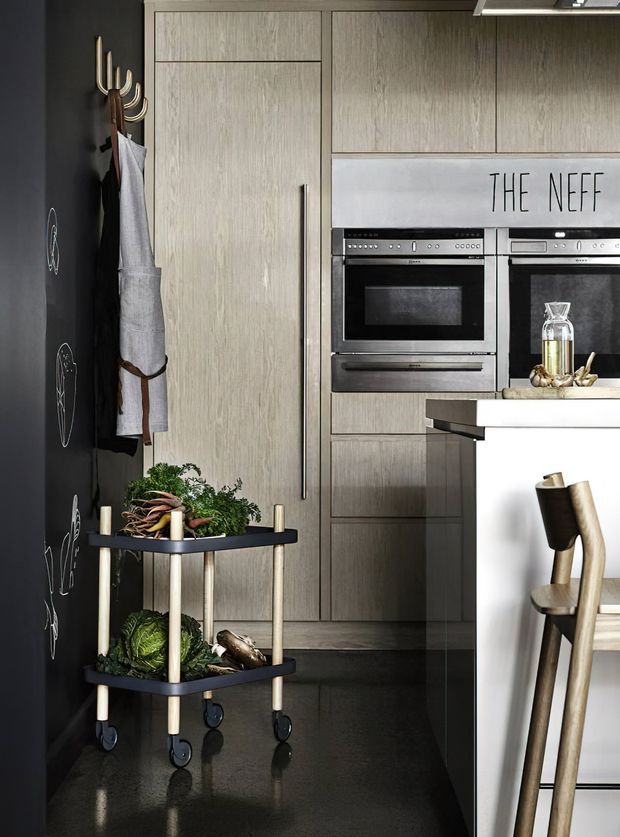 A compact contemporary kitchen with floor-to-ceiling pale timber cabinetry, integrated stainless-steel ovens, a white island, and a dark polished floor. A black feature wall with chalk-style drawings, hanging utensils, and a small wheeled produce trolley adds contrast and personality.