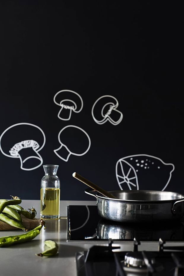 Close-up kitchen scene featuring a stainless-steel frying pan on a black glass cooktop, a glass bottle of cooking oil, and vegetables on a dark countertop. The black backsplash has playful white line drawings of mushrooms and other food-related forms, creating a bold, graphic backdrop.
