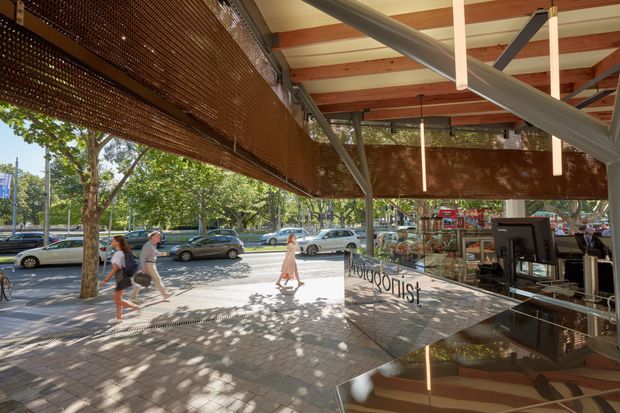 A bright, contemporary commercial interior viewed from a glazed storefront, with a café or retail counter and computer terminal on the right. Large glass panels reflect the exposed timber ceiling structure and angled steel beams, while woven timber or bamboo screening creates a warm textured canopy and partial enclosure. A tree-lined urban footpath and street are visible outside.