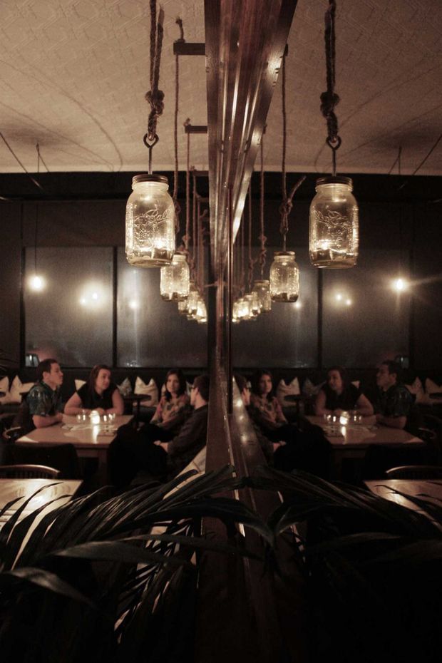 A dimly lit restaurant or hospitality interior with intimate dining tables, banquette seating, dark walls, and rows of hanging mason-jar pendant lights suspended from rough ropes. A large central mirrored or reflective structural feature creates strong symmetry and reflections, while dark leafy planting occupies the foreground. The atmosphere is warm, moody, and industrial-rustic.