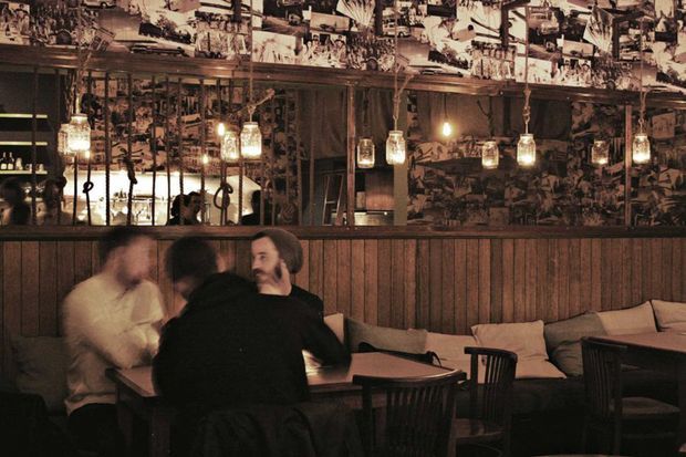A dimly lit, rustic restaurant or bar interior with dark timber wall panelling, a long counter with shelving and bottles, hanging glass pendant lights, and collage-style black-and-white imagery covering the upper walls and ceiling. Wooden tables and chairs with cushioned bench seating occupy the foreground, with patrons seated at one table. The atmosphere is intimate, warm, and moody.