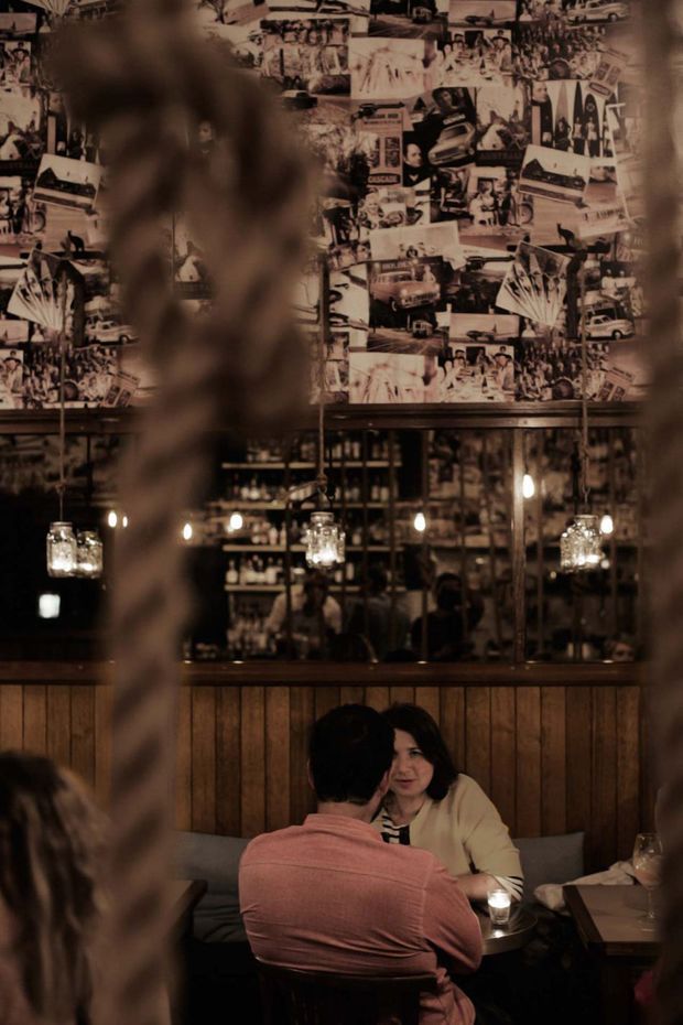 A dimly lit, intimate restaurant or bar interior viewed through blurred foreground elements. The space features a dark timber-paneled wall, a large mirrored bar-back with bottles and shelving, hanging glass pendant lights, and a collage-style vintage black-and-white photographic wall covering. Diners sit at a small table in the foreground. The atmosphere is warm, moody, eclectic, and vintage-inspired.