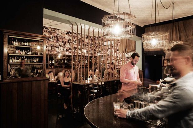 A dimly lit, richly styled bar or restaurant interior with a curved stone or dark composite counter in the foreground, a backlit drinks cabinet, hanging wire and glass pendant lights, dark walls, and a decorative partition made from timber poles and woven or rope-like elements. Several patrons and bartenders occupy the space, creating a warm, intimate hospitality atmosphere.