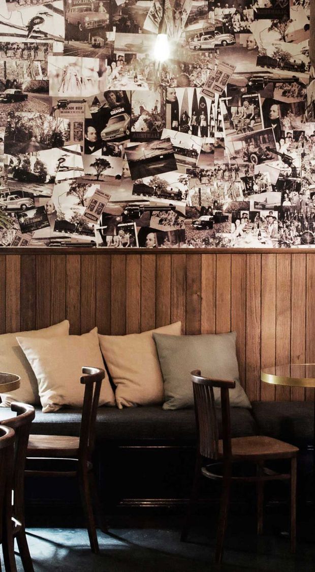 A moody, vintage-inspired seating nook featuring a dark upholstered banquette, wooden dining chairs, neutral cushions, and a wall covered with a dense collage of sepia historical photographs. Vertical dark timber wall panelling forms the backdrop, with warm low lighting and strong shadows creating an intimate atmosphere.
