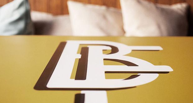 Close-up of a mustard-yellow tabletop or sign surface featuring a large bold white and dark-brown geometric graphic or lettering. A softly blurred upholstered seating area with cream cushions and warm timber wall panelling is visible in the background, suggesting a styled interior or hospitality setting.
